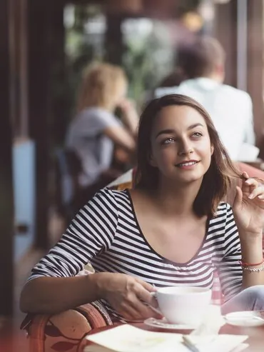Femeie la o masă de cafenea, cu o ceașcă de cafea, privind spre stradă